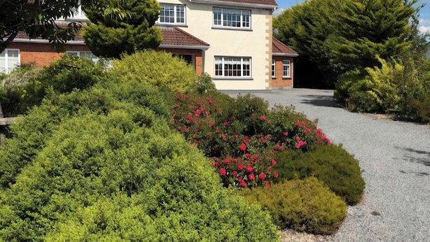 A two-story house with a gravel driveway and a garden with red flowers.