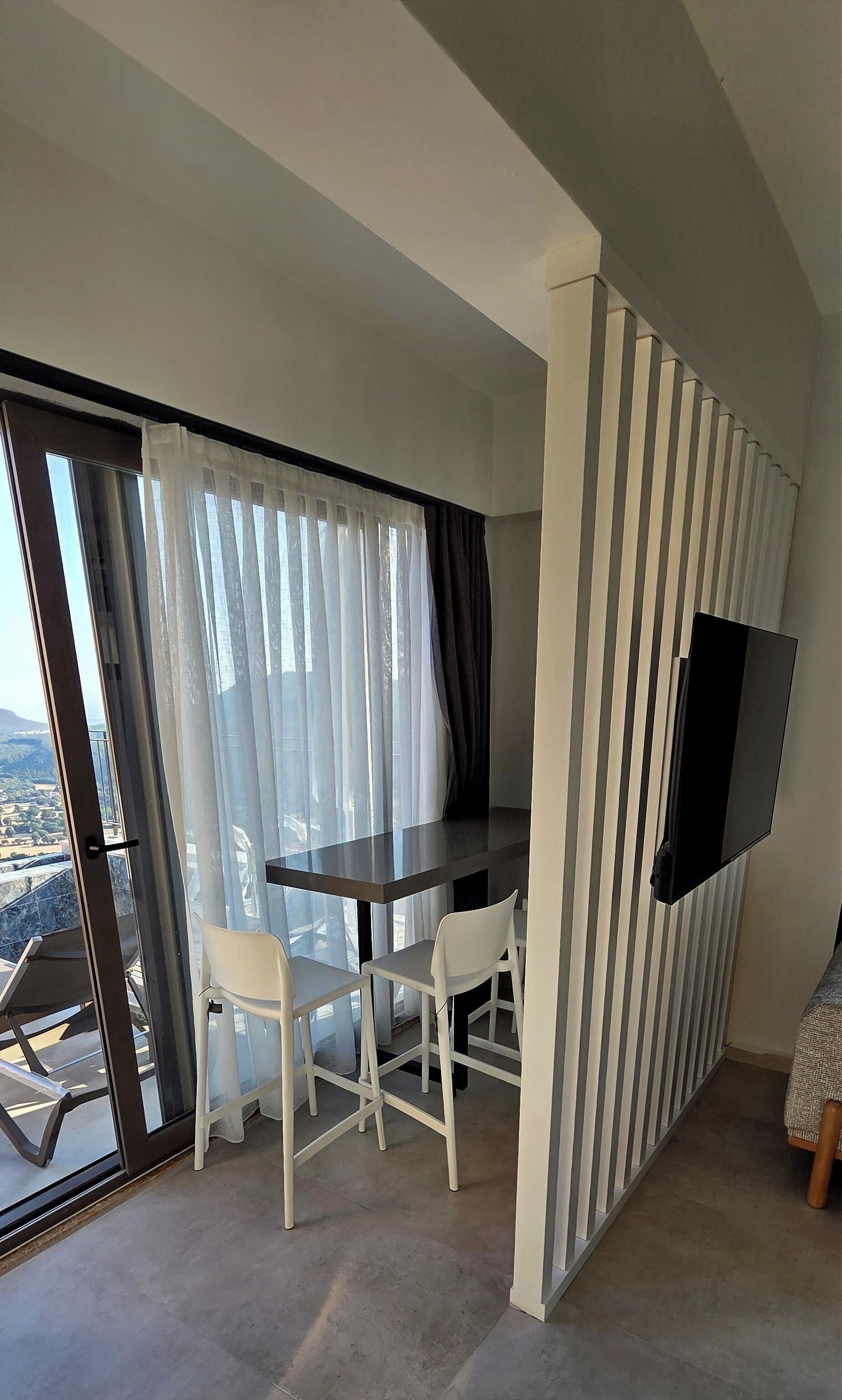 A modern room with a dining table, chairs, a TV mounted on the wall, and a large window with curtains.