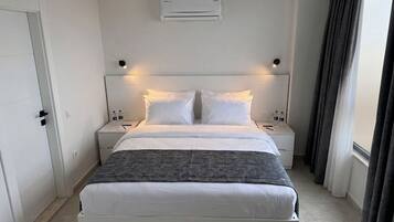 A modern hotel room with a bed, bedside tables, an air conditioning unit, and a window with curtains.