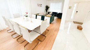 Een moderne eetruimte met een witte tafel en stoelen, een kroonluchter en uitzicht op een woonkamer met een groene bank.