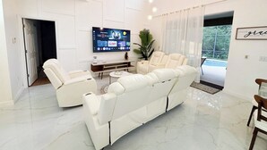 Een ruime woonkamer met een grote flatscreen-tv, een witte leren bank, een bijzettafeltje en een potplant.