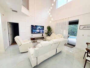 A spacious living room with a large flat-screen TV, a white leather sofa, a coffee table, and a potted plant. - Spacious 7-bedroom condo in terrific Kissimmee with AC (Kissimmee)