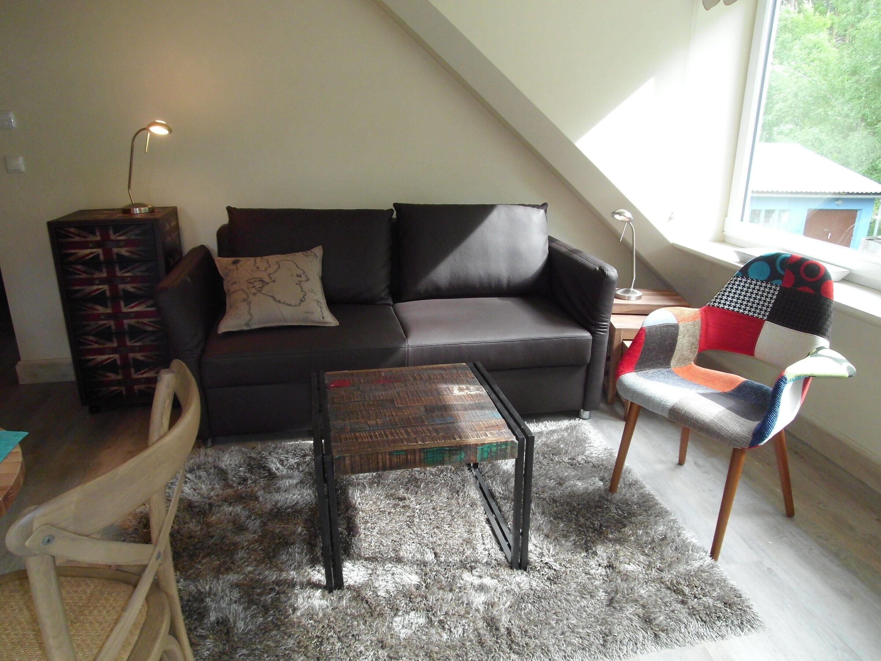 Una sala de estar tipo loft con un sofá oscuro, una mesa de centro de madera, un sillón estampado y una mesita con una lámpara.