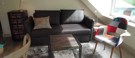Una sala de estar tipo loft con un sofá oscuro, una mesa de centro de madera, un sillón estampado y una mesita con una lámpara.