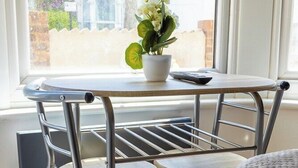 Ein Zimmer mit Fenster, einem Tisch mit Metallgestell und Stühlen, einer Topfpflanze und einer Fernbedienung.