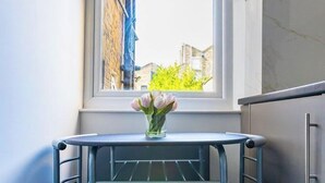 Ett litet kök med ett fönster, ett metallbord med två stolar och en vas med blommor.