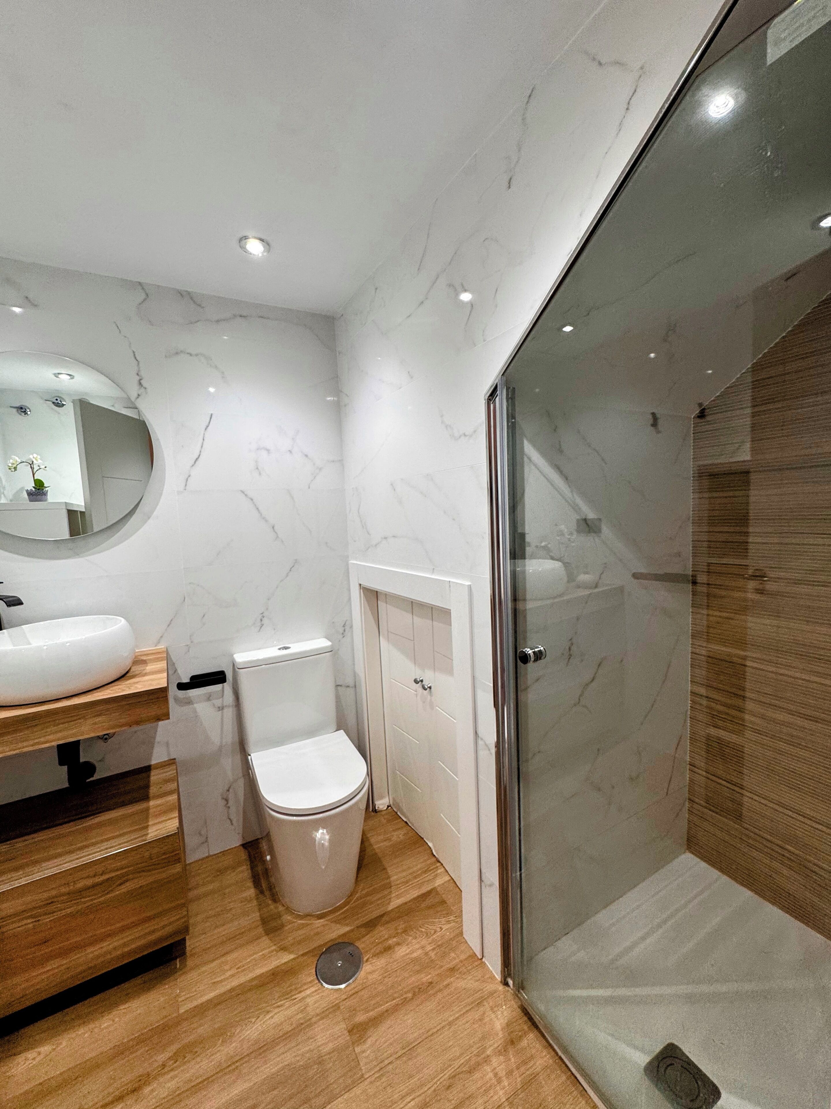 Een moderne badkamer met een marmeren wand, een houten wastafelmeubel, een toilet en een douchecabine met glazen wanden.