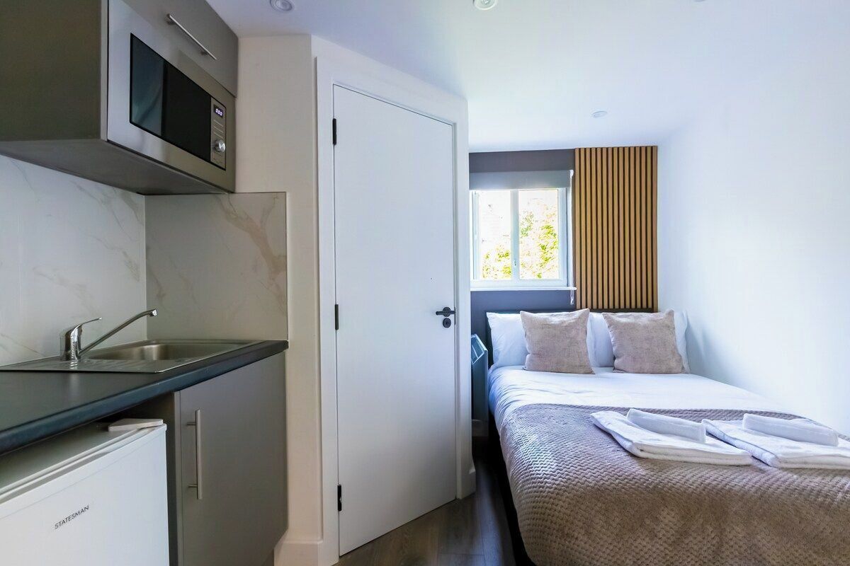 A compact hotel room with a kitchenette, a bed with a view, and a window with blinds.