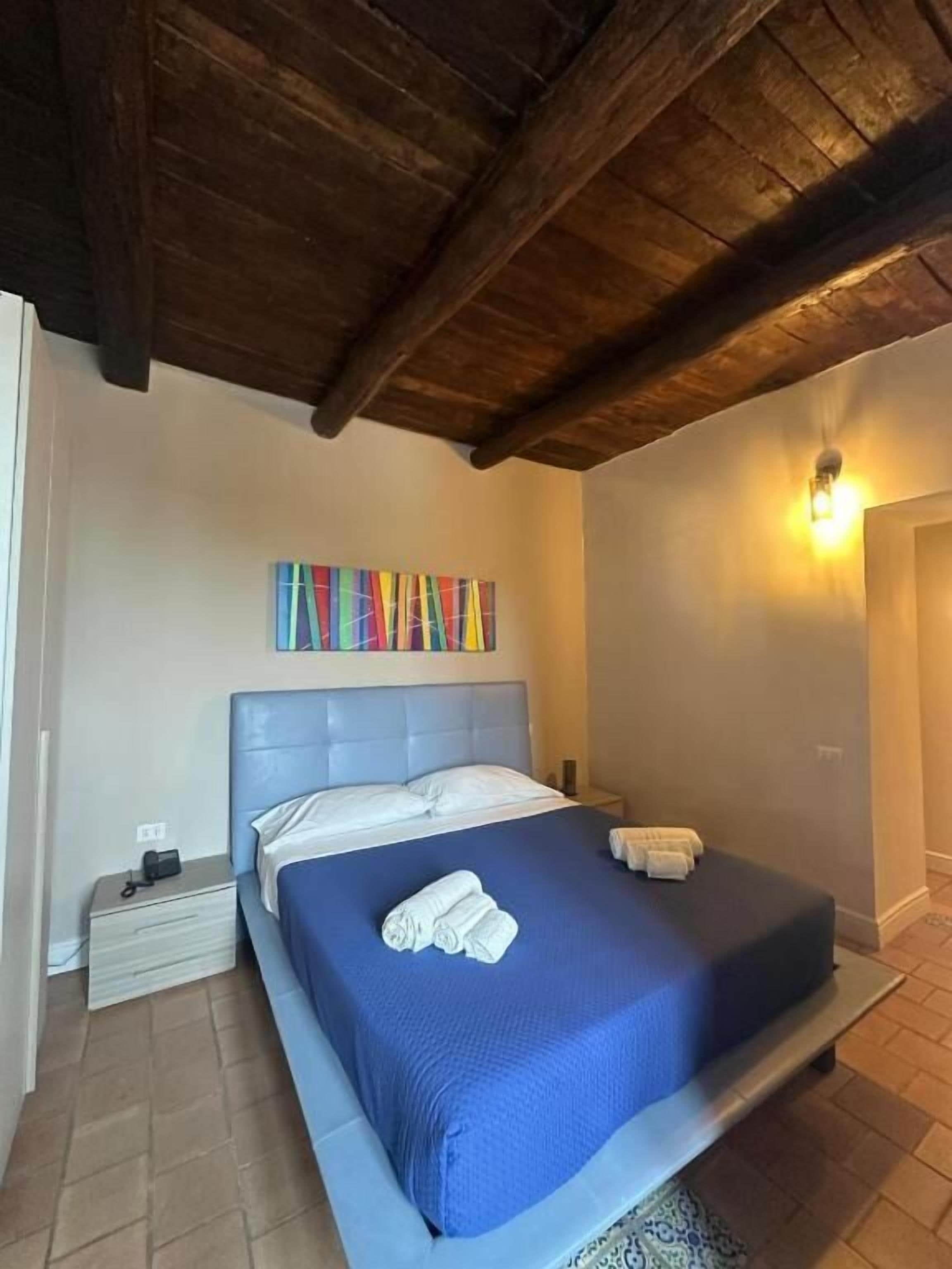 A bedroom with a wooden ceiling, a bed with a blue bedspread, a colorful artwork on the wall, and a nightstand with a lamp.