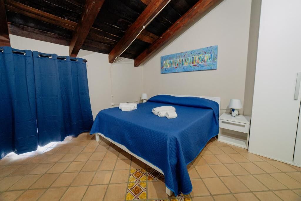 Ein Schlafzimmer mit einem Bett, einem Nachttisch, einer Lampe, einem Bild an der Wand und einem blauen Vorhang.