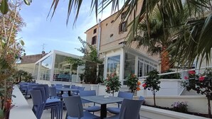 Un espacio al aire libre con mesas y sillas, rodeado de plantas y un edificio con cerramiento de vidrio.