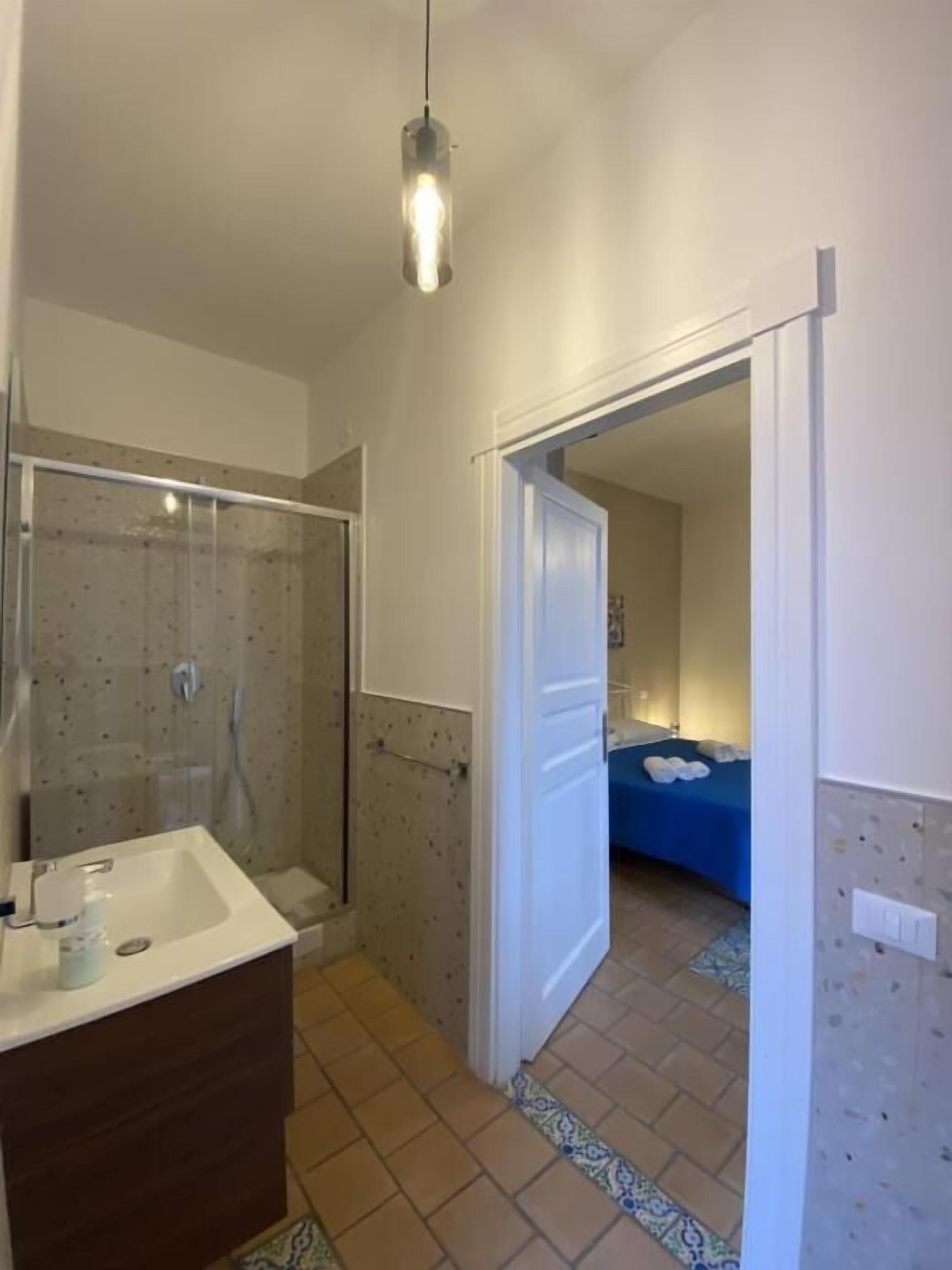 A bathroom with a shower, a sink, and a mirror. There is a door leading to a bedroom.
