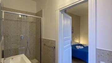 A bathroom with a shower, a sink, and a mirror. There is a door leading to a bedroom.