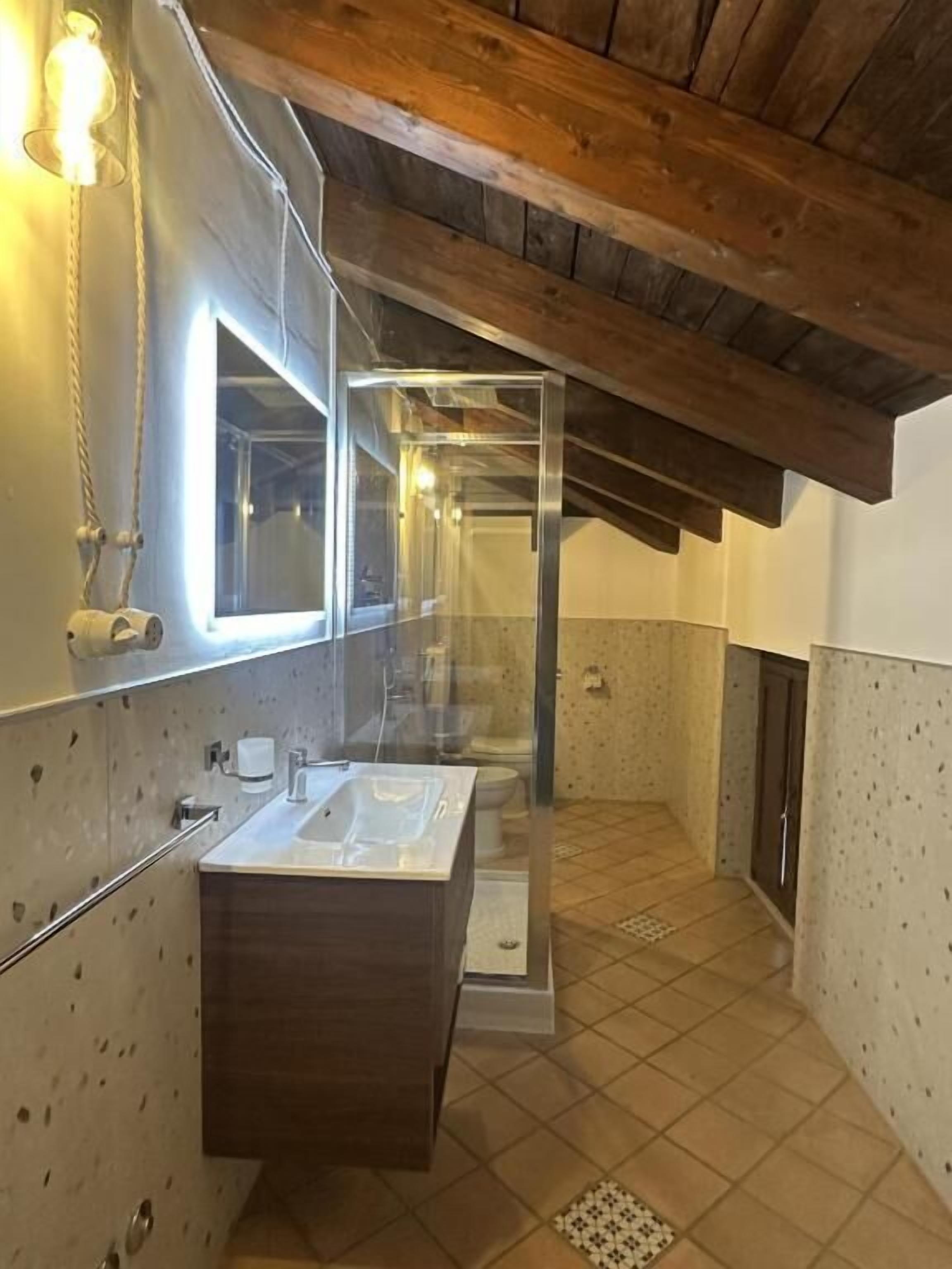 Een badkamer met een houten plafond, een glazen douchecabine, een wastafel met spiegel en betegelde vloer.