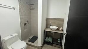 A bathroom with a toilet, a shower with a tiled wall, a sink with a wooden countertop, and a door. - Patriota Loft for 2, with amenities and parking (Monterrey)