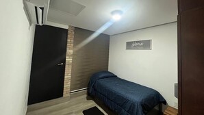 A small, single-bed room with a wooden floor, a black door, a wall-mounted air conditioning unit, a small bedside table, and a sign on the wall. - Patriota Loft for 2, with amenities and parking (Monterrey)