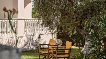 Uma área externa com assentos de madeira, cadeiras e uma mesa, rodeada por vegetação e um edifício com varandas.