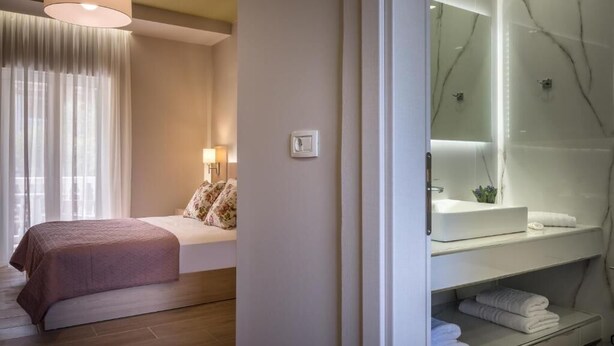 A bedroom with a bed, a bedside lamp, and a view of a bathroom with a sink and mirror. - Katerina Studios (Planos)