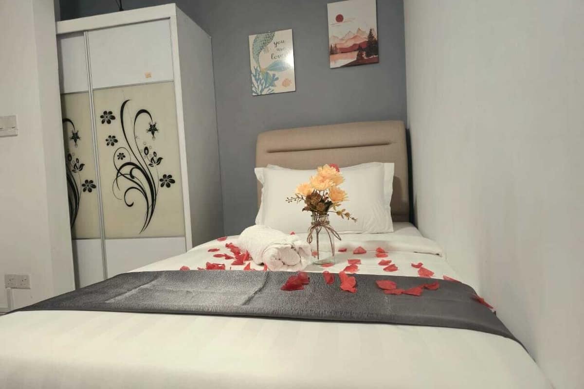 A neatly made bed with a white duvet, a grey throw blanket, and red rose petals scattered on it.