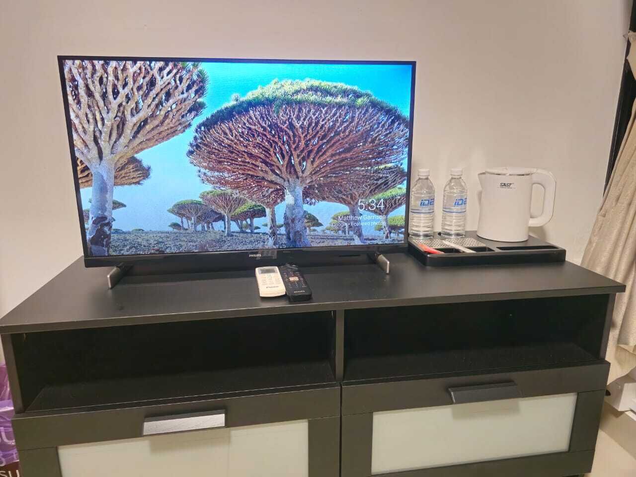 A television on a black entertainment unit with a remote control, a white mug, and two water bottles.