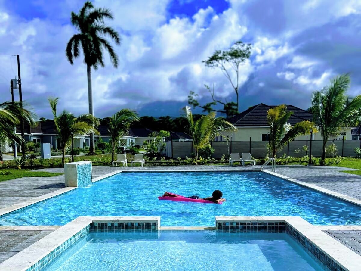 A swimming pool with a person floating on a pink float, surrounded by palm trees and a cloudy sky.