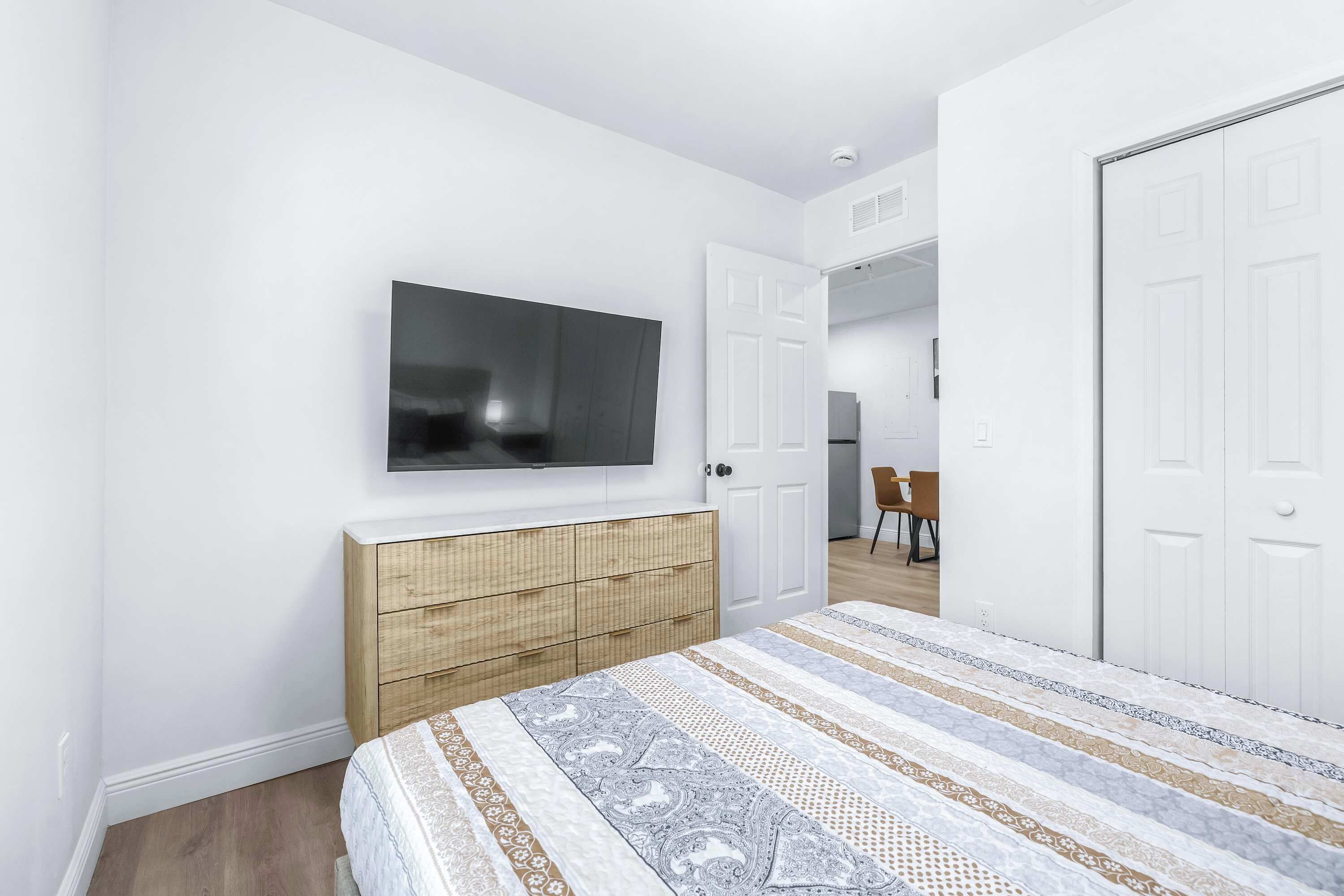 A bedroom with a bed, a TV mounted on the wall, a wooden dresser, and white doors.