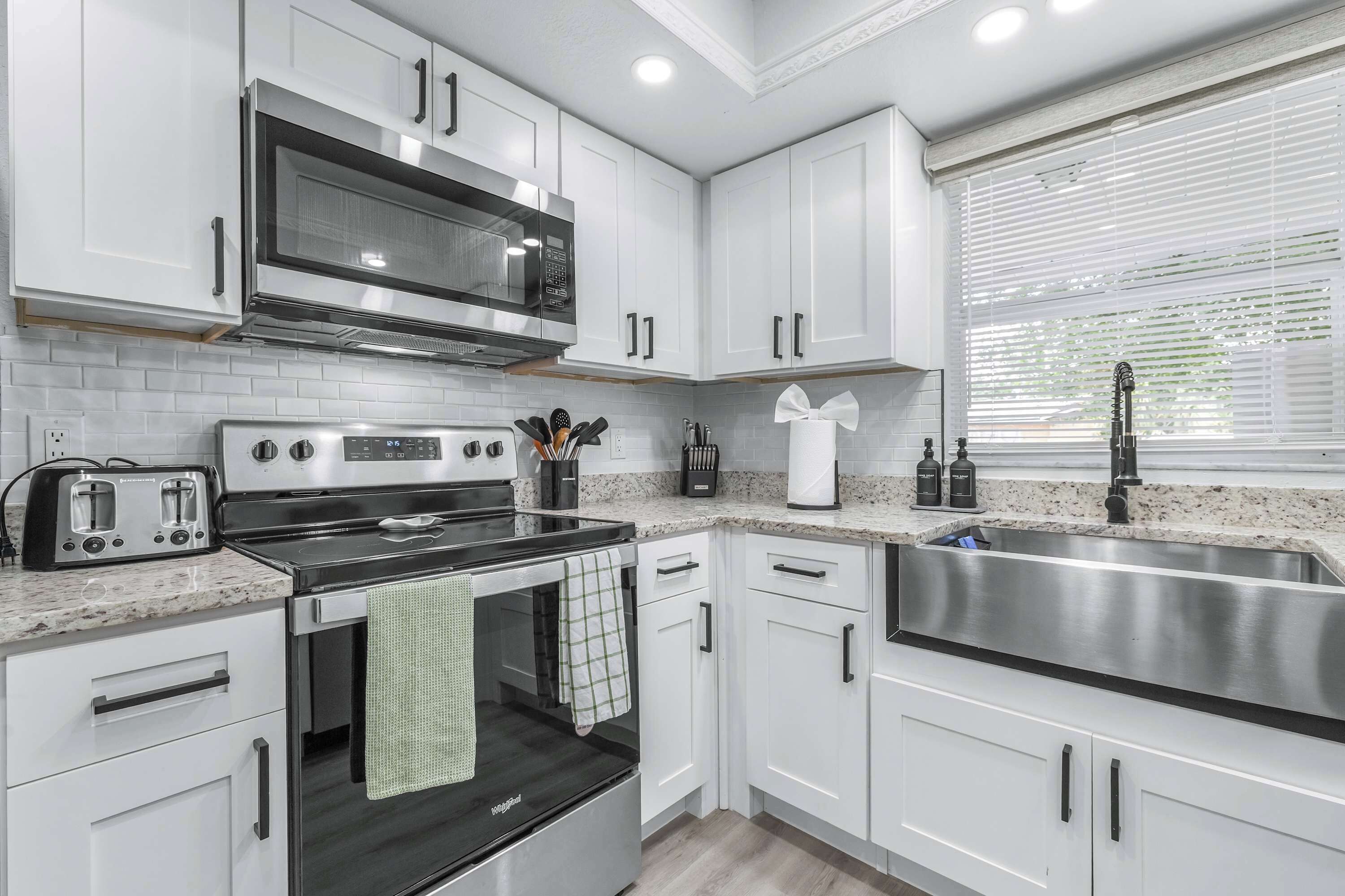 Une cuisine moderne avec des meubles blancs, un évier en acier inoxydable, un four à micro-ondes, une cuisinière et une fenêtre avec des stores.