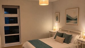 A bedroom with a bed, bedside tables, a hanging light fixture, a window, and a painting on the wall. - Spacious 2 Bed apartment Rainham LDN Gated Parking (Greater London)