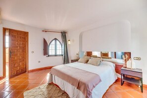 Ein Schlafzimmer mit Bett, Nachttisch, einem Fenster mit Vorhängen, einer Holztür und einem Fliesenboden.