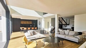 Un salon moderne avec un canapé, une table basse et des fauteuils. On aperçoit un coin cuisine au fond.