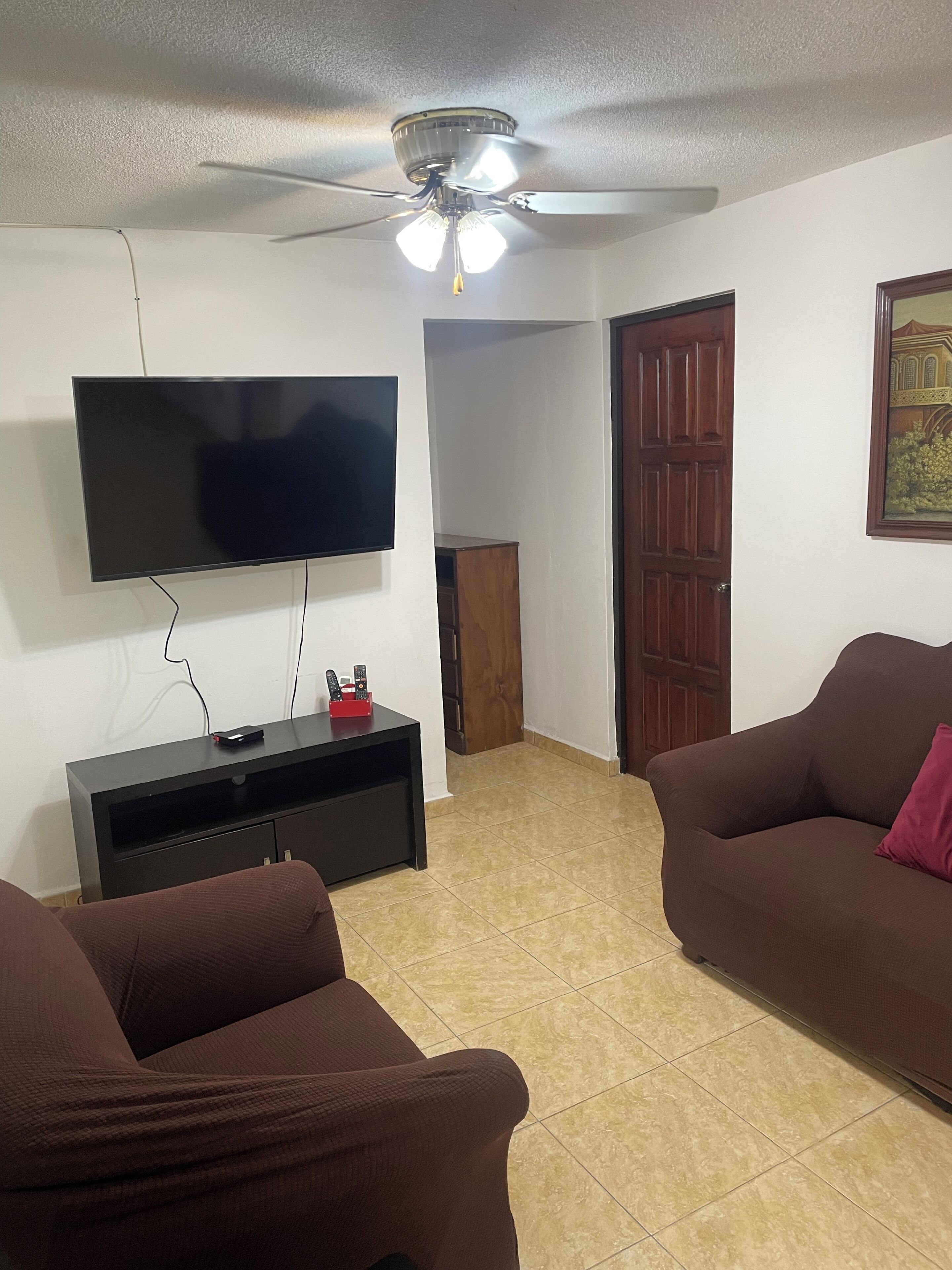 A living room with a flat-screen TV, a ceiling fan, two brown sofas, a wooden door, and a tiled floor.