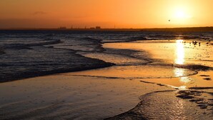 Ein Strand mit Wellen, einem Sonnenuntergang und einer entfernten Stadtlinie am Horizont.