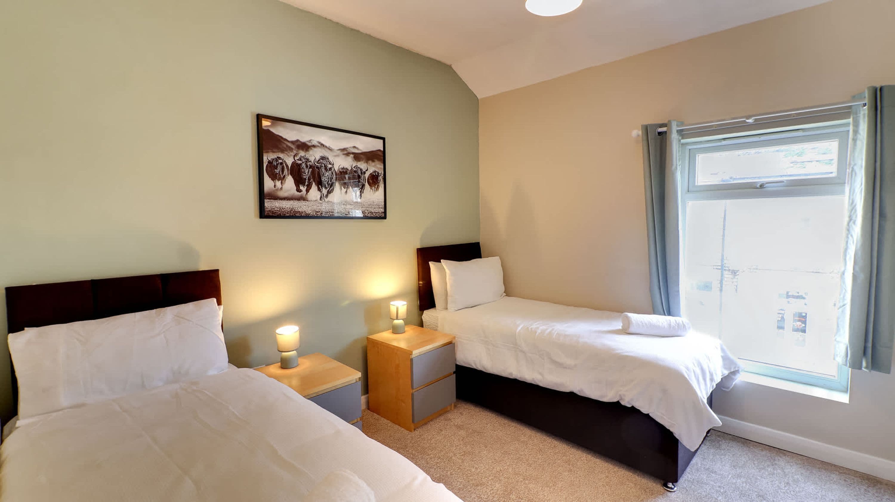 A hotel room with two beds, a nightstand with a lamp, a framed picture on the wall, and a window with curtains.