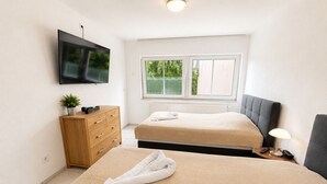 Un dormitorio con cama, cómoda, televisor, ventana y mesita de noche con lámpara.