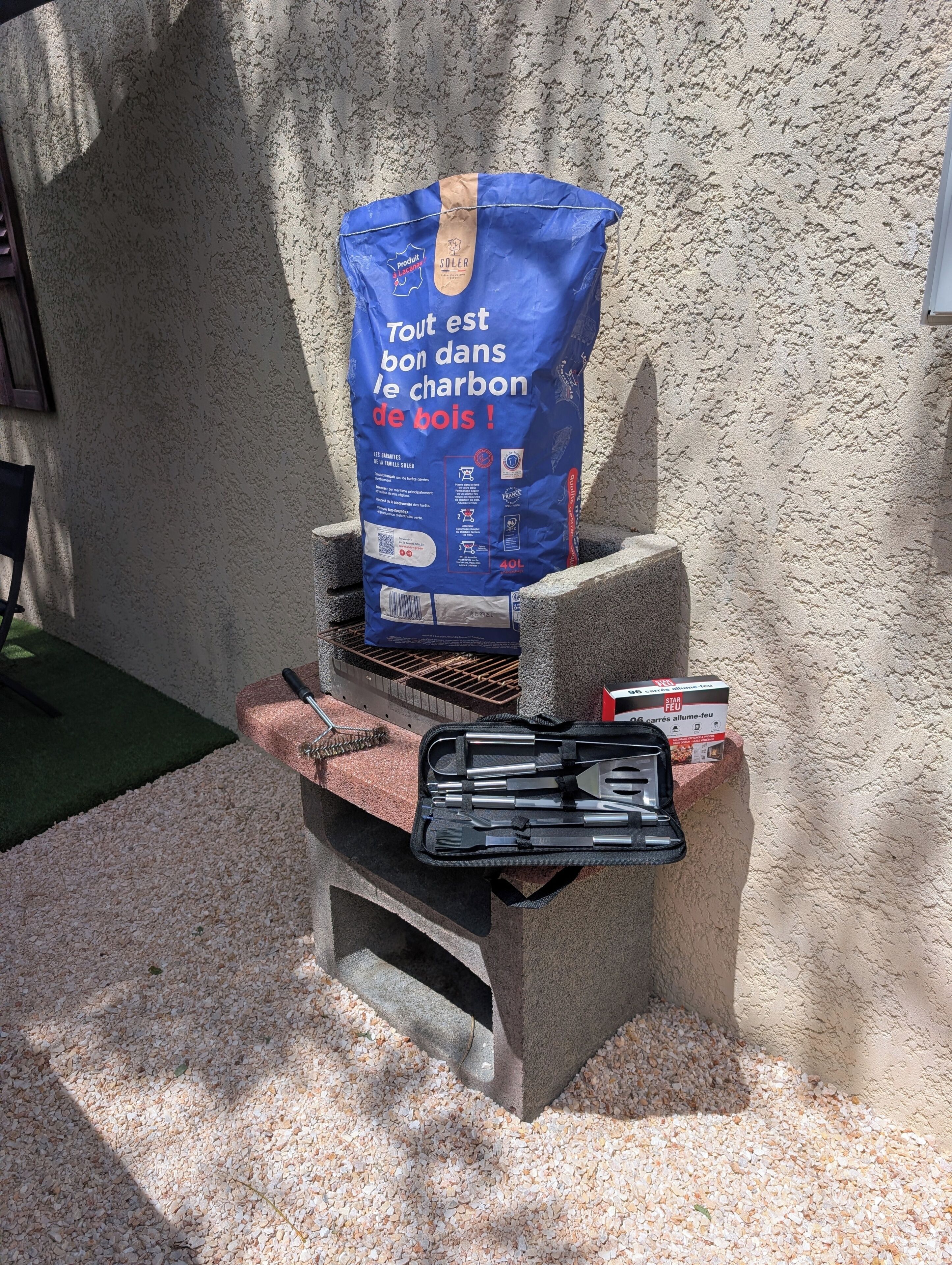 Un barbecue au charbon de bois, avec un sac de charbon et les ustensiles nécessaires pour griller.
