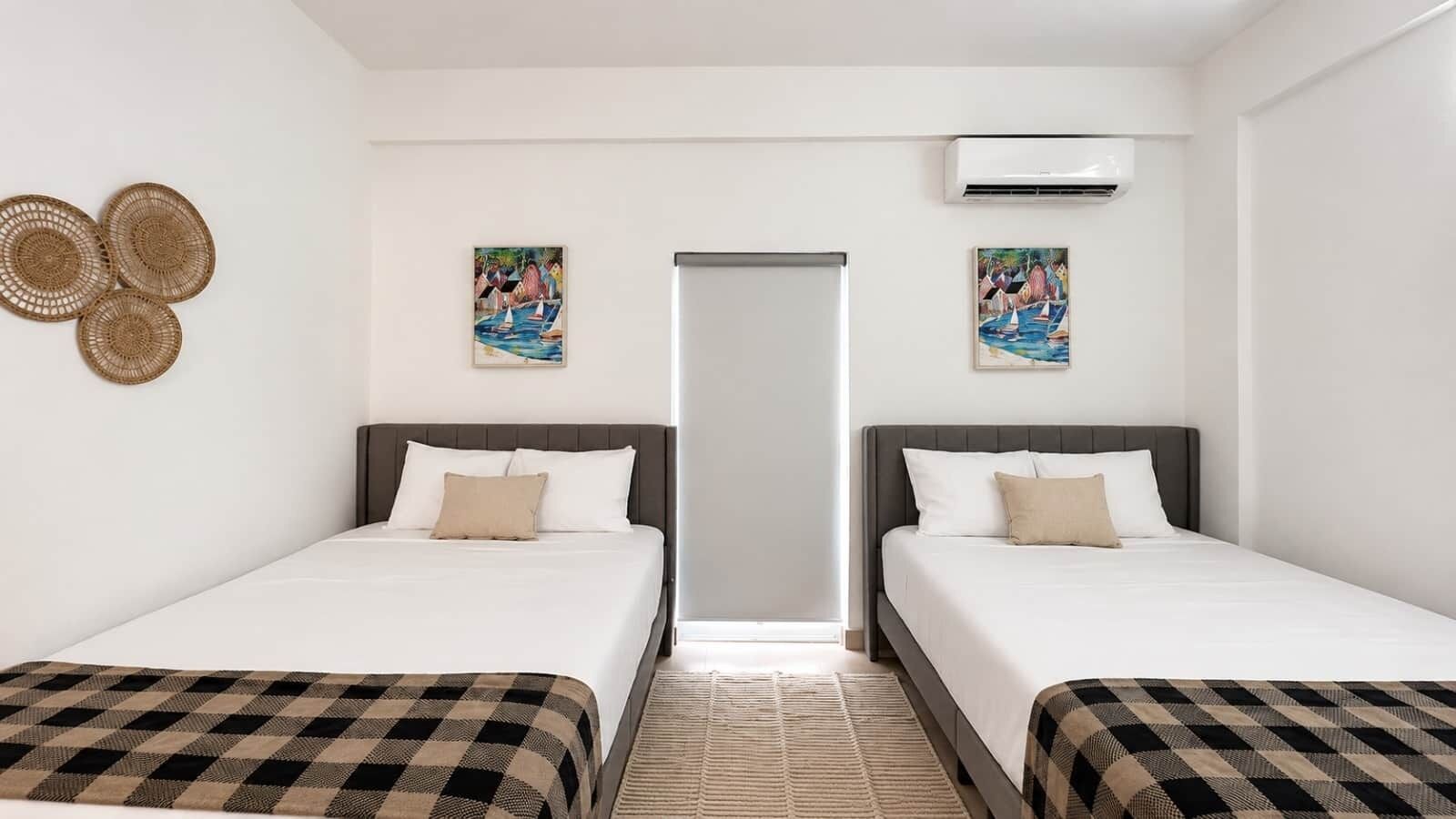 Ein Hotelzimmer mit zwei Betten, einer karierten Tagesdecke, einer gewebten Wanddekoration und zwei Bildern an den Wänden.