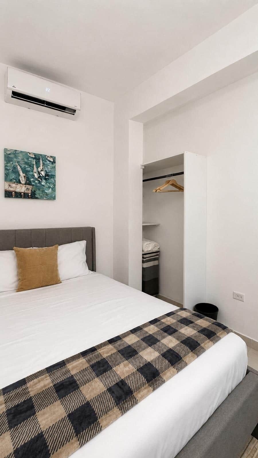 A hotel room with a bed, a painting, a wardrobe, and an air conditioning unit.