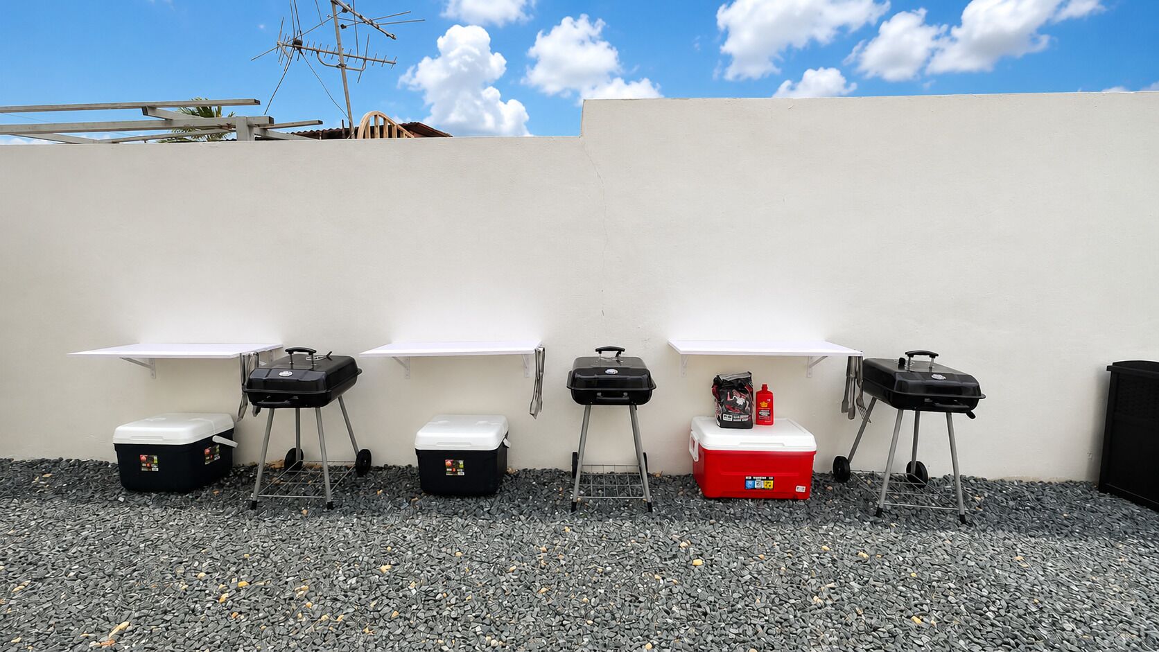 Parrillas para barbacoa al aire libre, enfriadores y un enfriador rojo con una botella sobre una superficie de grava frente a una pared blanca.