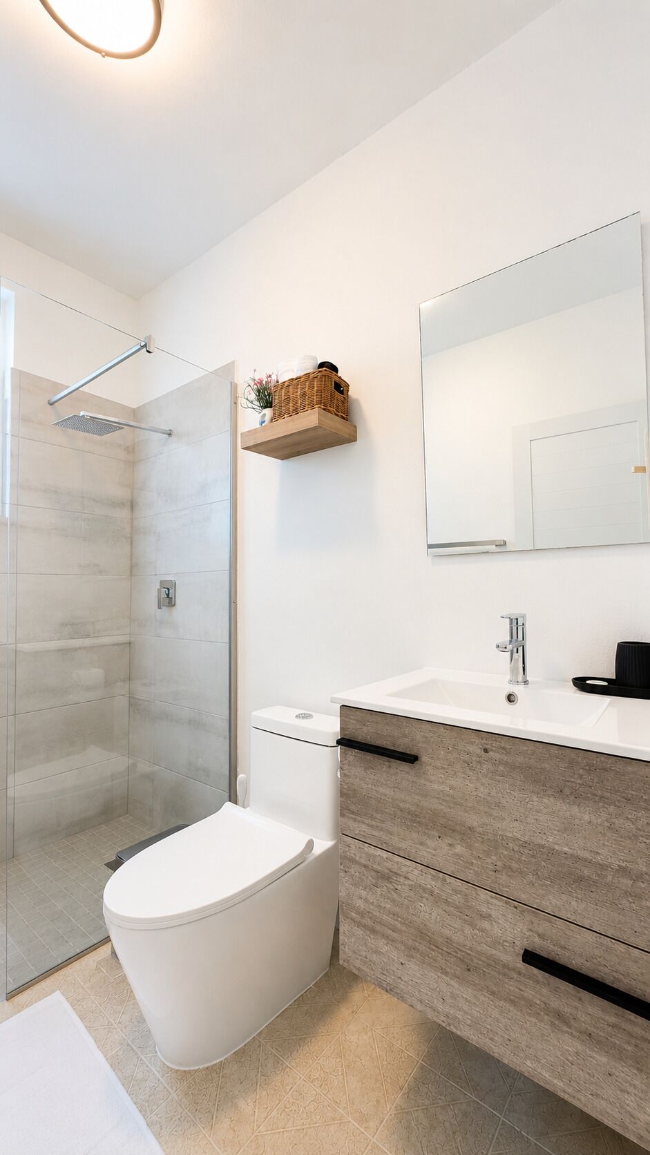 A modern bathroom with a glass shower enclosure, a toilet, a vanity with a mirror, and a small shelf with toiletries.