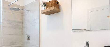 A modern bathroom with a glass shower enclosure, a toilet, a vanity with a mirror, and a small shelf with toiletries.