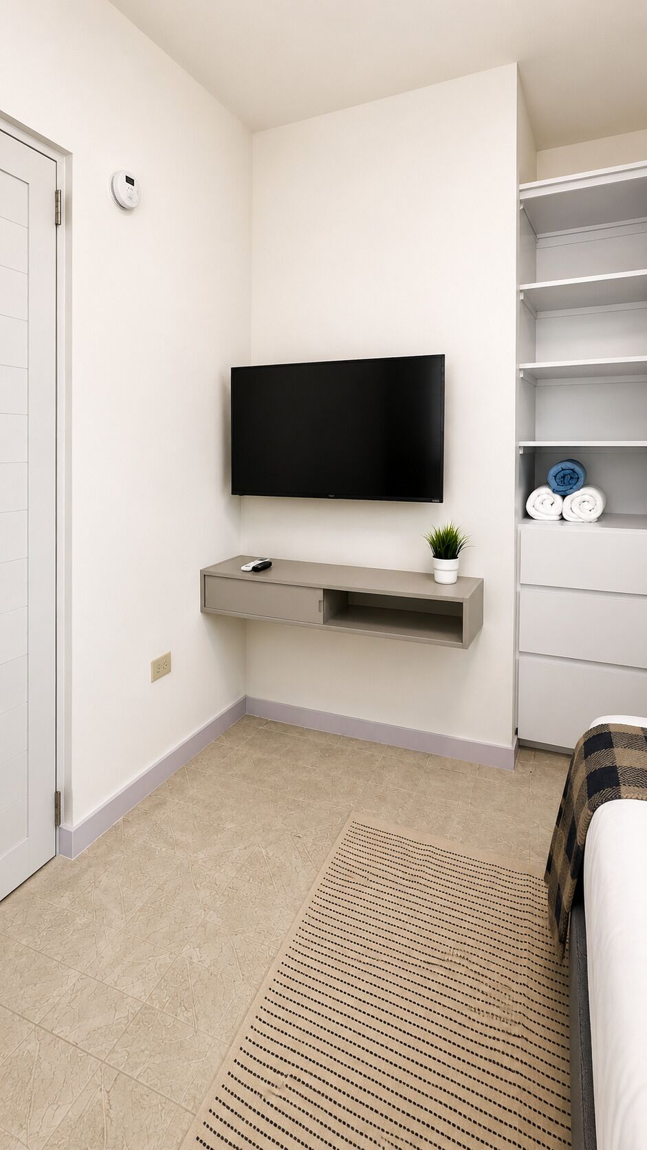 Une chambre à coucher avec un téléviseur à écran plat fixé au mur, une table de chevet avec une télécommande, une petite plante et un lit recouvert d’une couverture à carreaux.