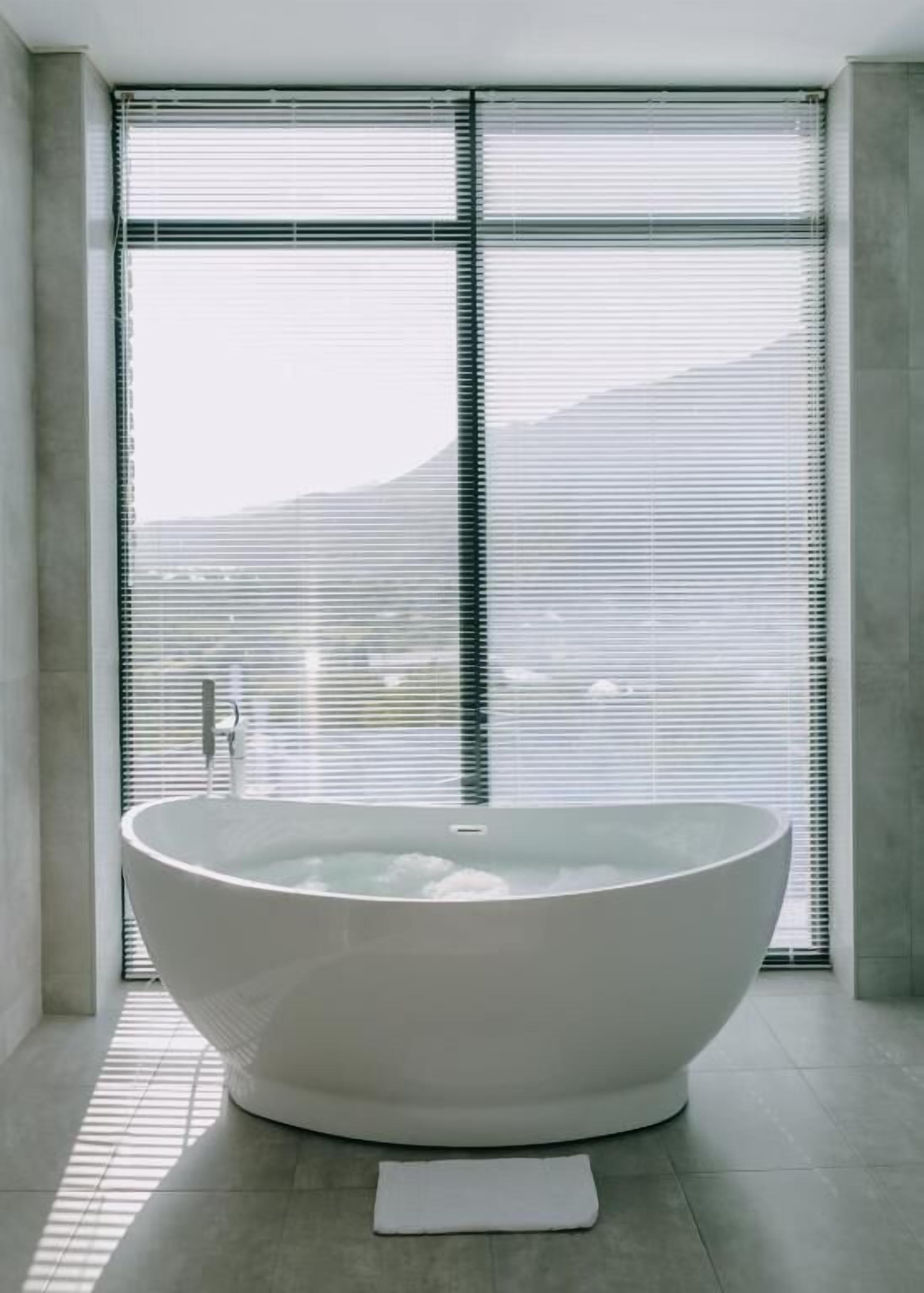 A modern bathroom with a large freestanding bathtub, large windows with blinds, and a view of a mountain.