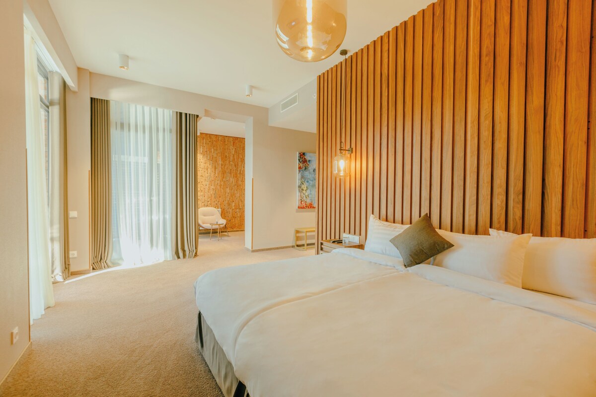 A hotel room with a large bed, a wooden accent wall, a painting, and a view of the outdoors.