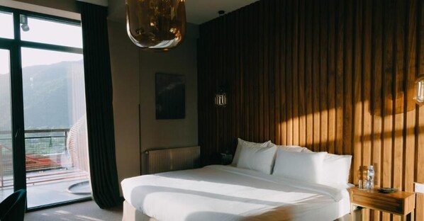 A hotel room with a large bed, a wooden wall panel, a window with a view, and a hanging light fixture.