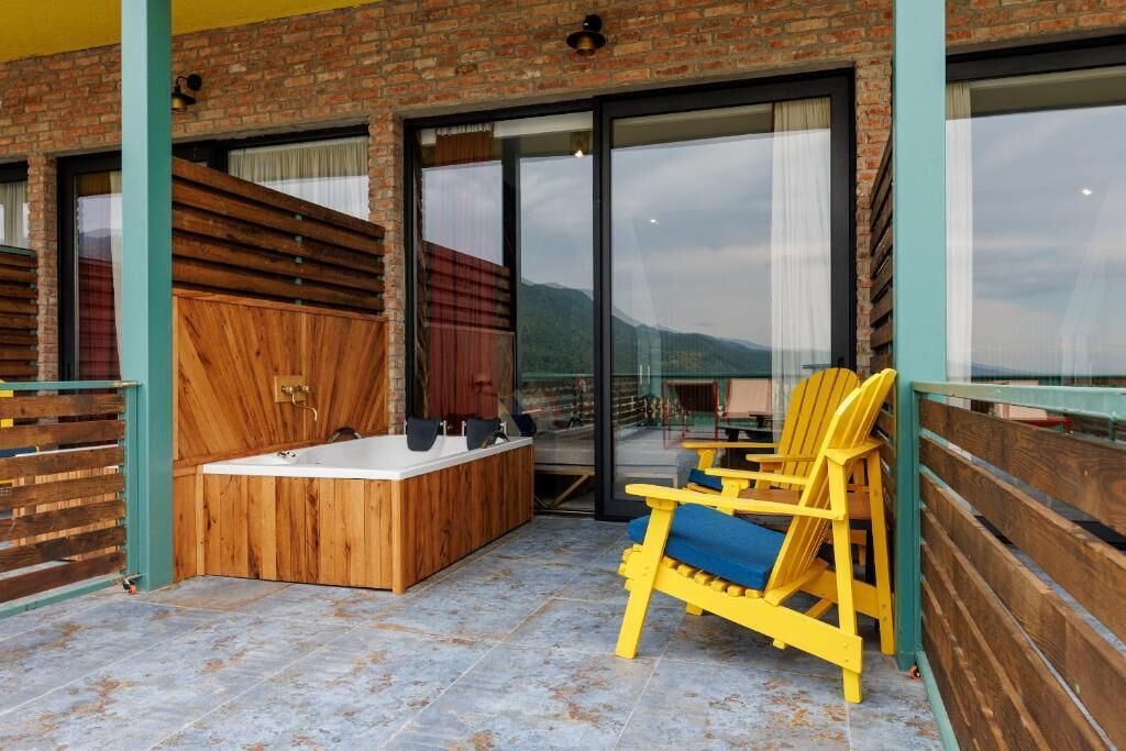 Uma área de spa com uma banheira de madeira, uma cadeira amarela e uma porta de vidro que dá para uma varanda com vista para as montanhas.