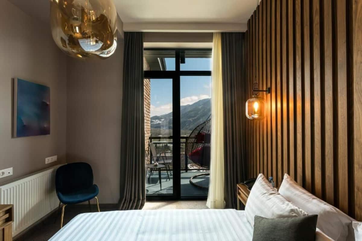 A hotel room with a bed, a chair, a balcony with a view of mountains, and a hanging light fixture.