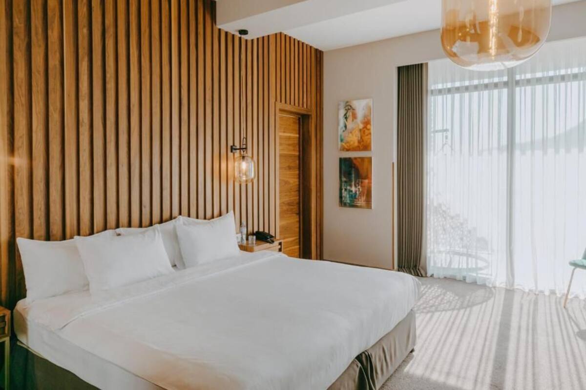 A hotel room with a large bed, wooden panel wall, a hanging light fixture, and a view through the window.