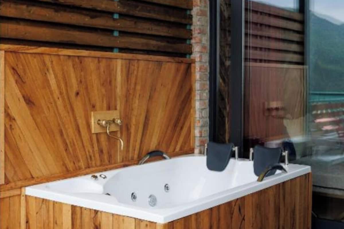 A wooden spa tub with a faucet and two jets, set against a brick wall and large window.
