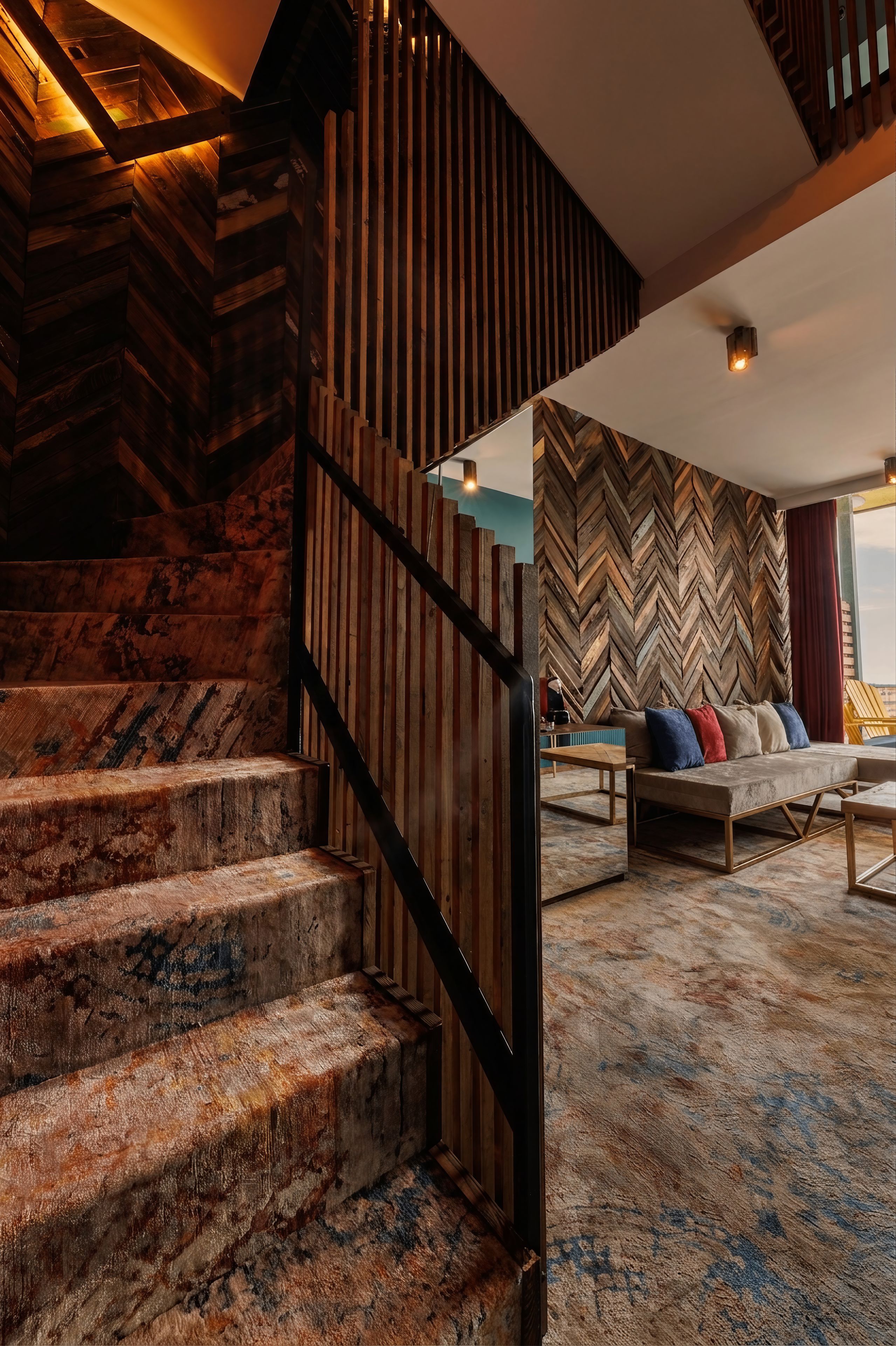 Un escalier avec des marches en bois et une rampe en métal, menant à une pièce dont un mur est décoré d’un motif en chevrons et qui contient un canapé avec des coussins.