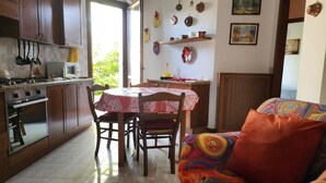 Uma cozinha com armários de madeira, uma mesa de jantar com cadeiras, um sofá com uma almofada colorida e uma janela com vista para o verde.